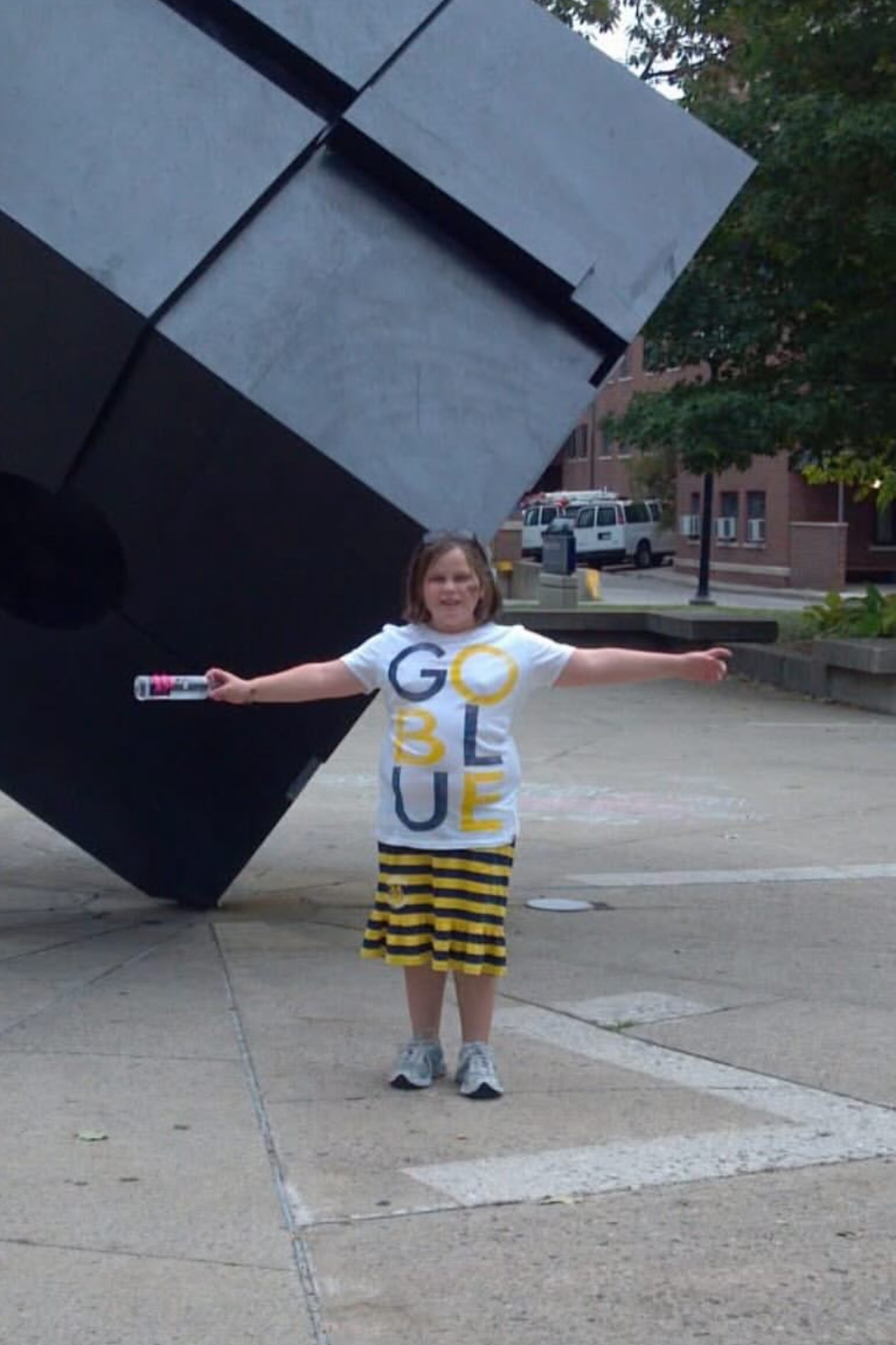 Grace as a child in a Go Blue t-shirt, arms outstretched in front of the Cube sculpture in Ann Arbor
