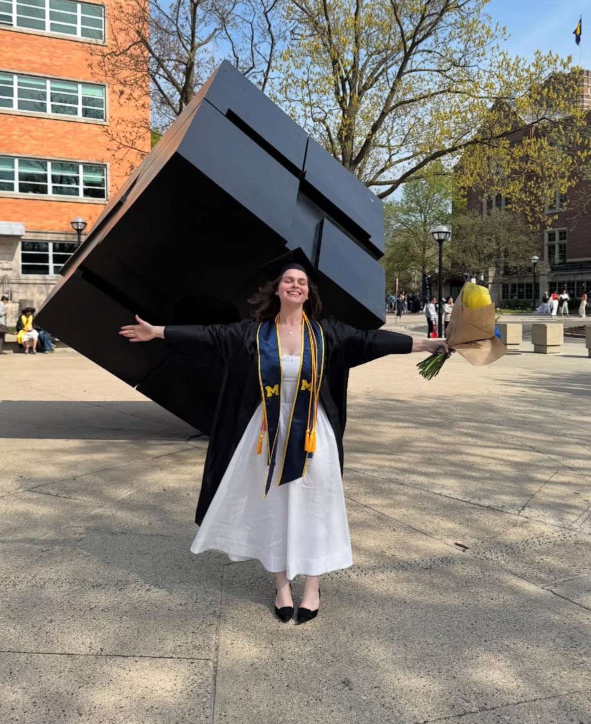 Grace in her cap and gown, arms outstretched in the same pose in front of the Cube, May 2026